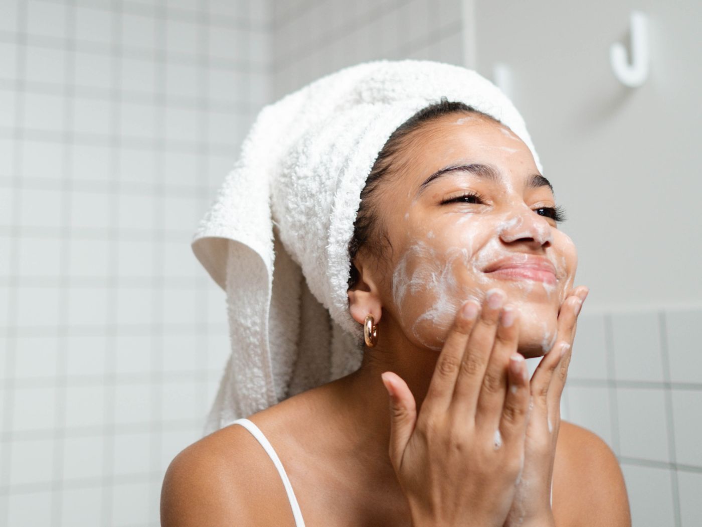 Woman in tiled bathroom exfoliating her face with a towel around her head