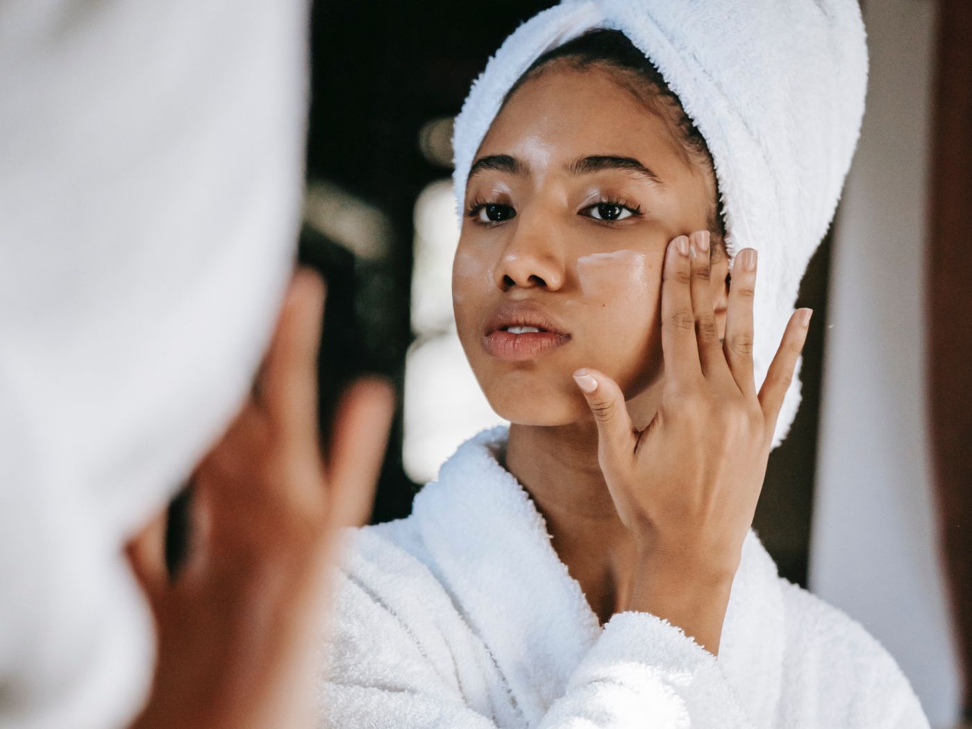 Woman in a towel hair wrap and robe putting moisturizer on her cheek