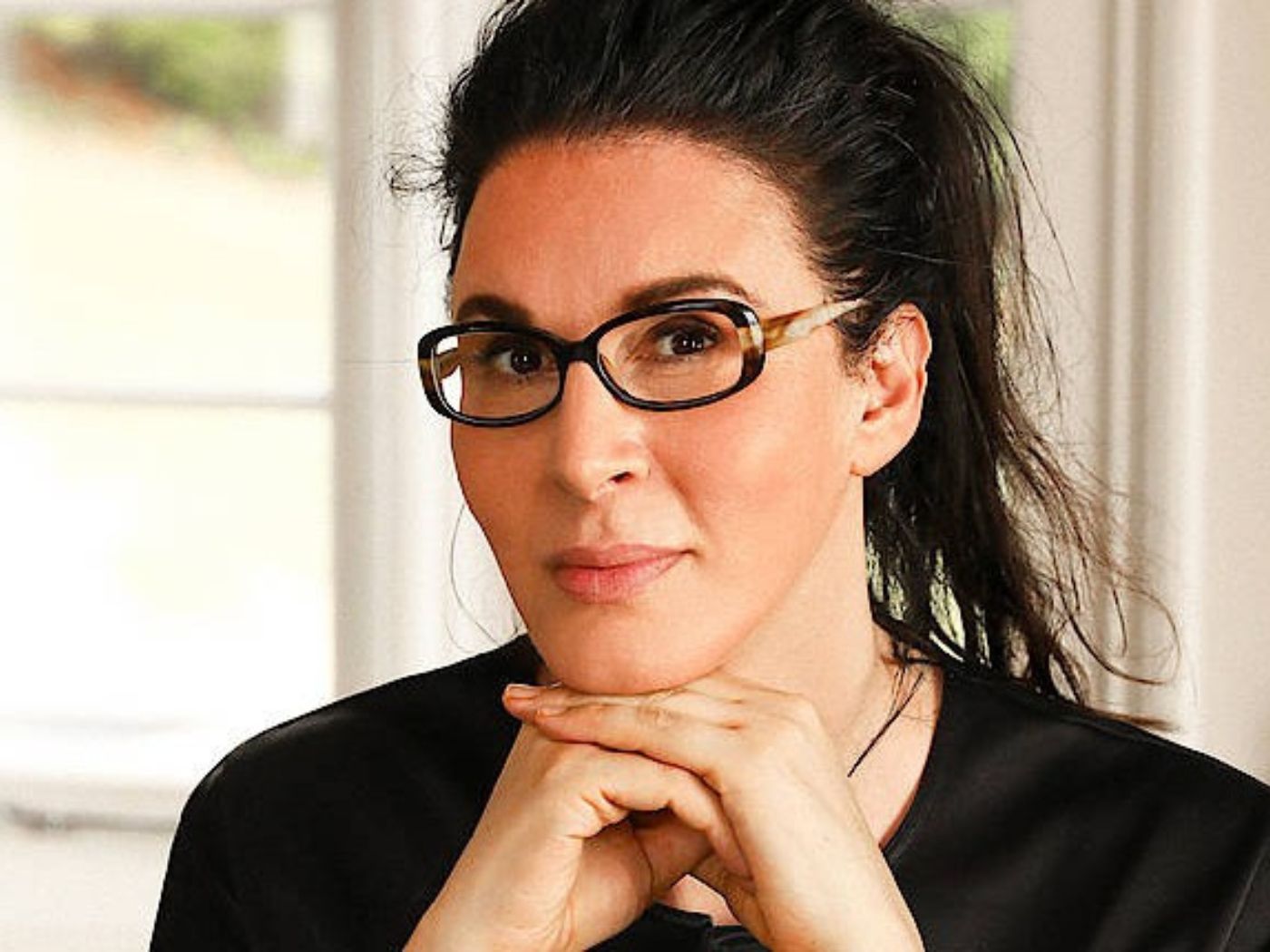 Woman wearing tortoiseshell glasses seated at desk, in front of a window.