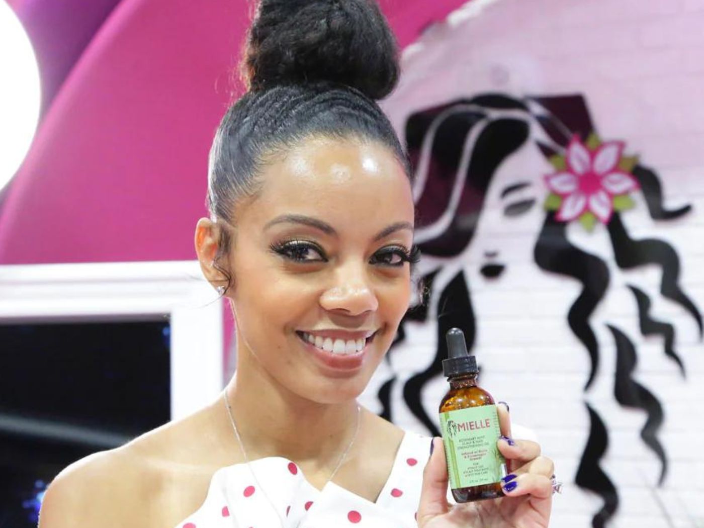 Monique Rodriguez stands smiling, holding a Mielle Organics product in a brown apothecary bottle, with a green label.